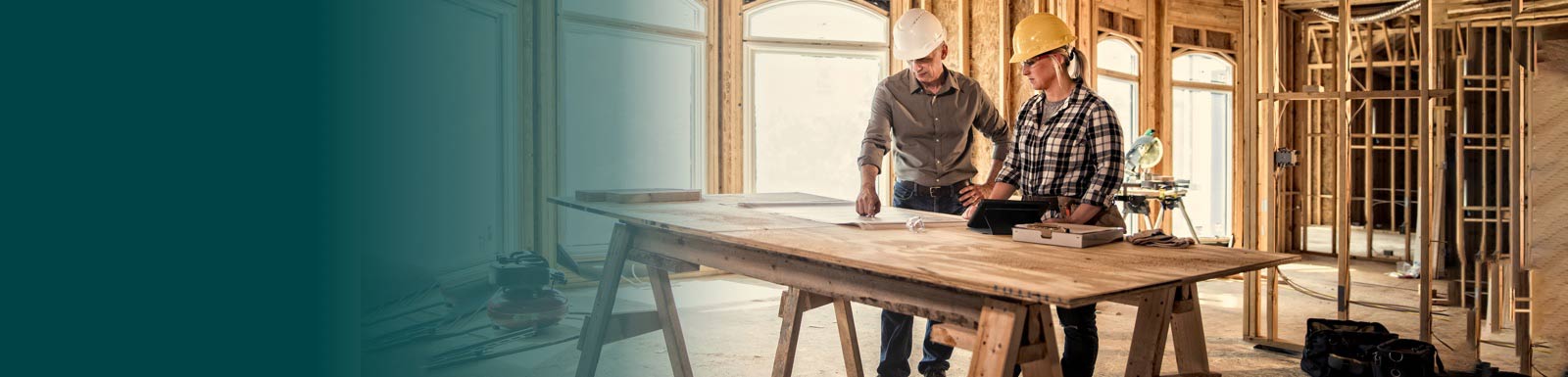 Two people looking at plans amid the wooden frame of a house being built